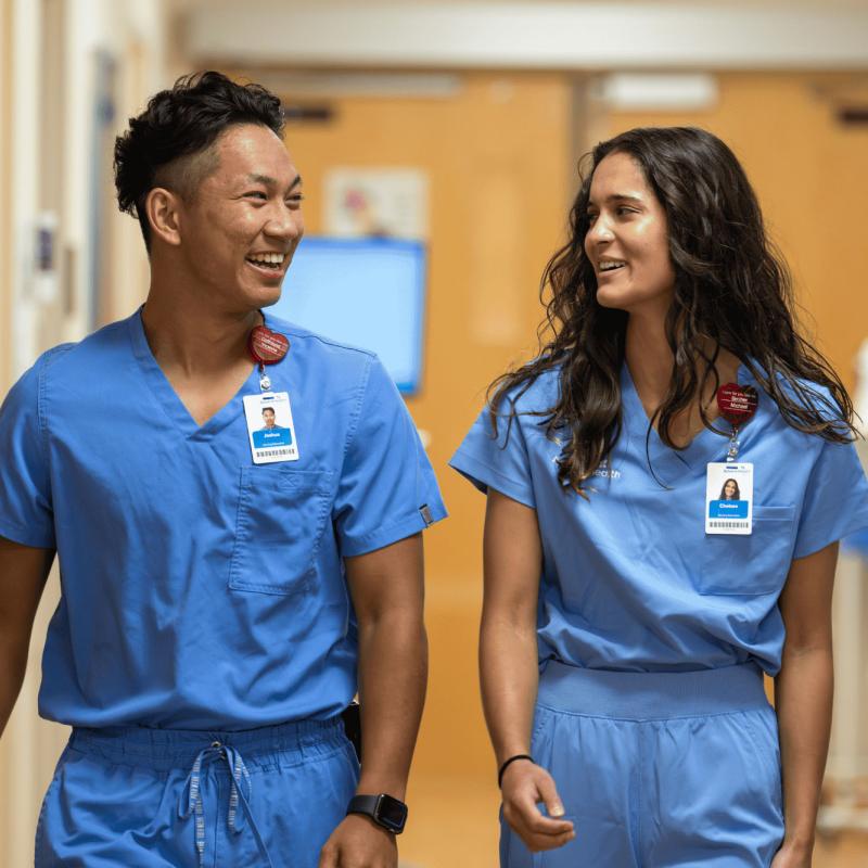A young male and young female nurse walk down a hospital hallway together